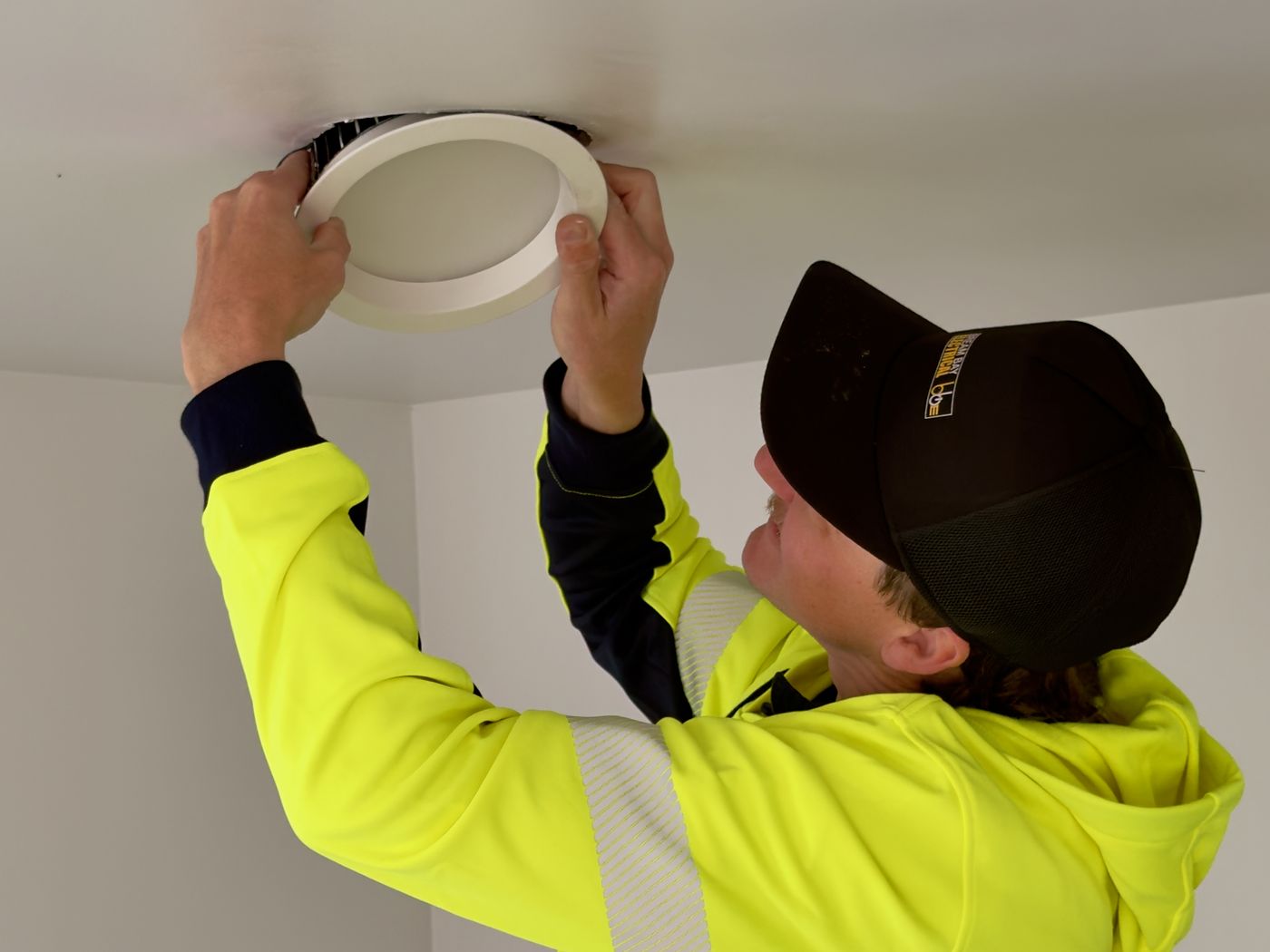 A Bream Bay Electrical team member in a hi-vis hoodie and black cap fitting a round white downlight into a ceiling