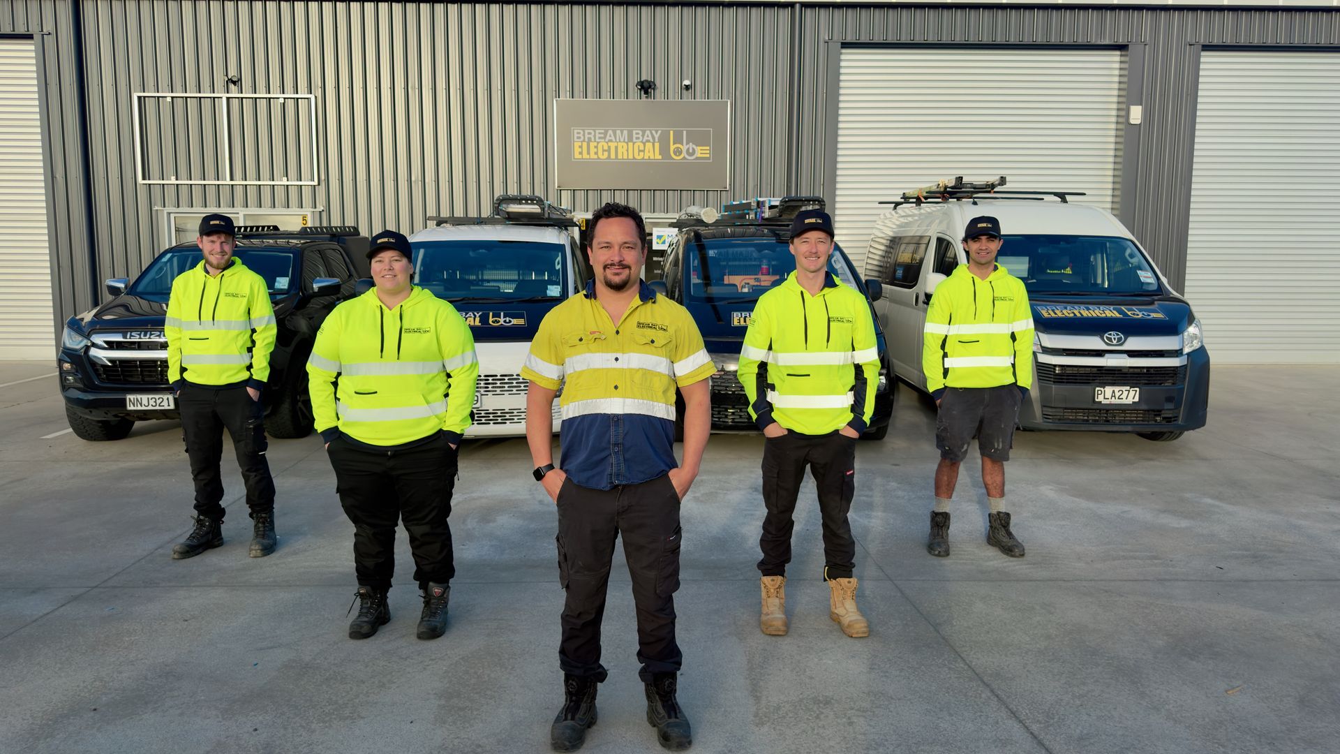 Five of the Bream Bay Electrical team in hi-vis yellow standing in front of four company vehicles outside the workshop, with the company sign on the wall behind them