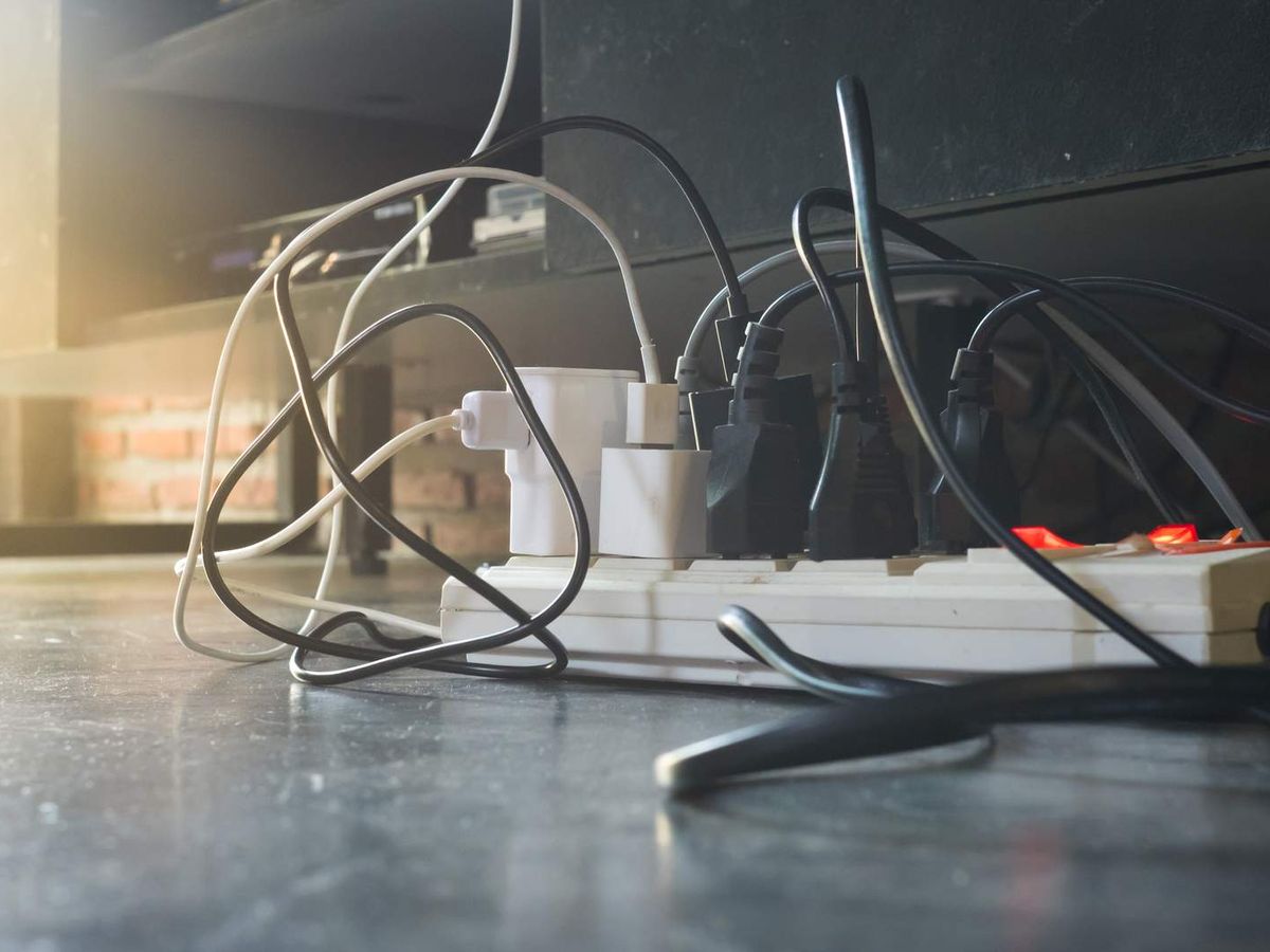 Plugs and adaptors crowded into a white power board on the floor, with black and white leads tangled around it