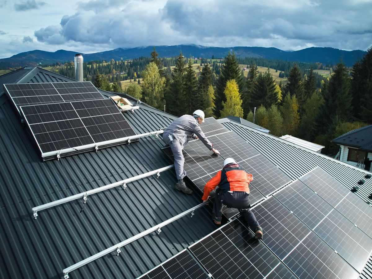 Two installers fixing solar panels to a dark metal roof under an overcast sky