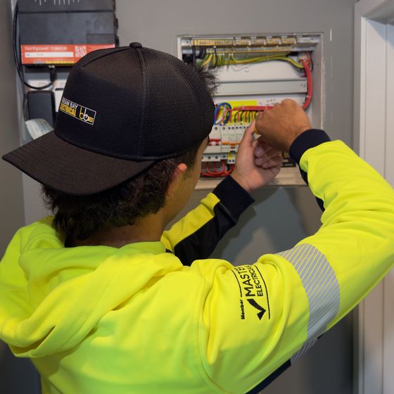 Seen from behind, a team member in a hi-vis hoodie and black cap with both hands on the wiring of an open switchboard